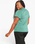 A woman wearing a heathered sage green short-sleeve t-shirt and black pants, shown from a side-back angle against a plain white studio background.