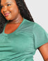 A close-up of a woman wearing a heather green short-sleeve V-neck t-shirt, accessorized with a gold chain necklace and hoop earrings, against a light gray background.