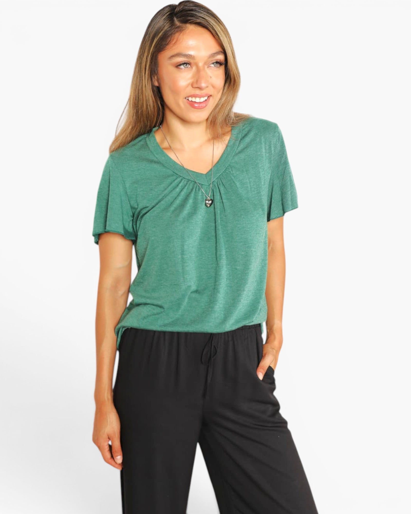 A woman wearing a green short-sleeve V-neck top with a gathered front detail and black drawstring pants, photographed against a plain light gray studio background.