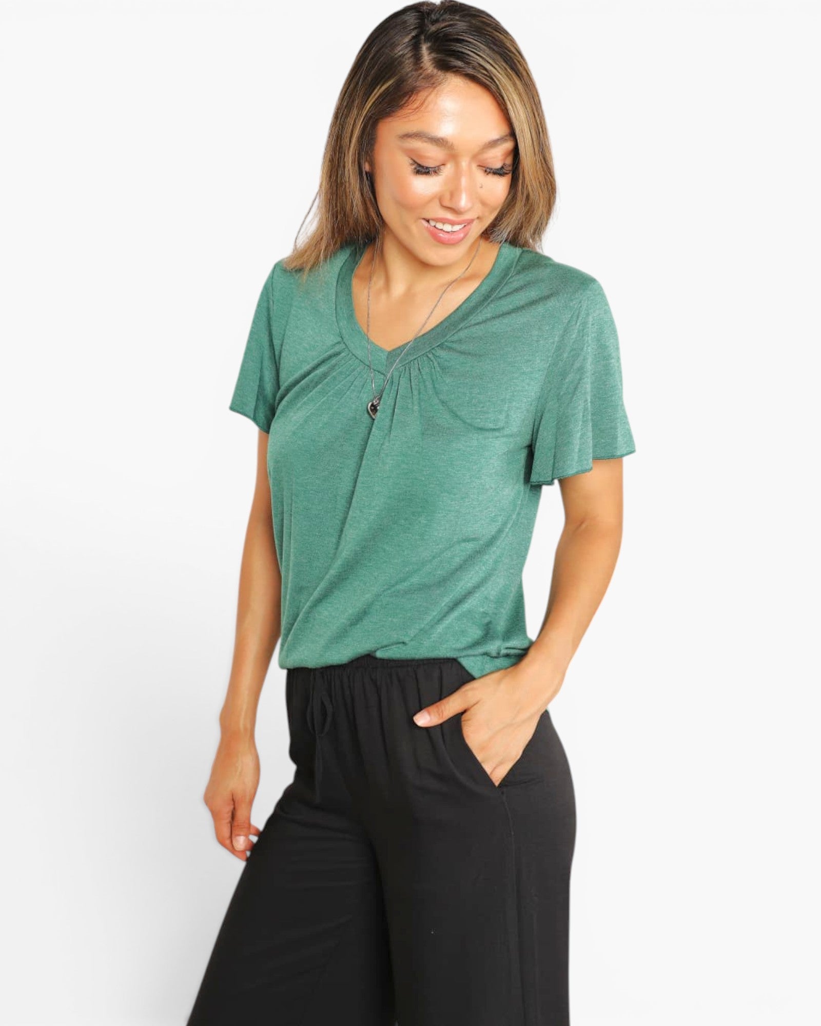 A woman wearing a heather green short-sleeve V-neck top with a chest pocket and black pants, standing against a light gray studio background.