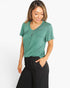 A woman wearing a heather green short-sleeve V-neck top with a chest pocket and black pants, standing against a light gray studio background.