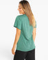 A woman with long blonde-highlighted hair is shown from the back wearing a heather green short-sleeve athletic top and black pants against a plain white studio background.