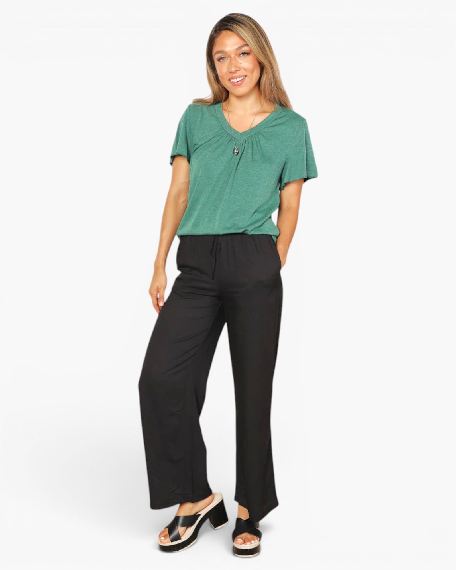 A woman modeling a short-sleeve green V-neck top with a small button detail and black wide-leg pants, standing against a plain white studio background.