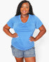 A woman modeling a heather blue short-sleeve V-neck t-shirt with a gathered chest detail, paired with denim shorts, against a plain light gray background.