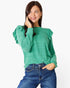 A woman wearing a green long-sleeve top with ruffled shoulders and blue jeans, posing against a light gray studio background.