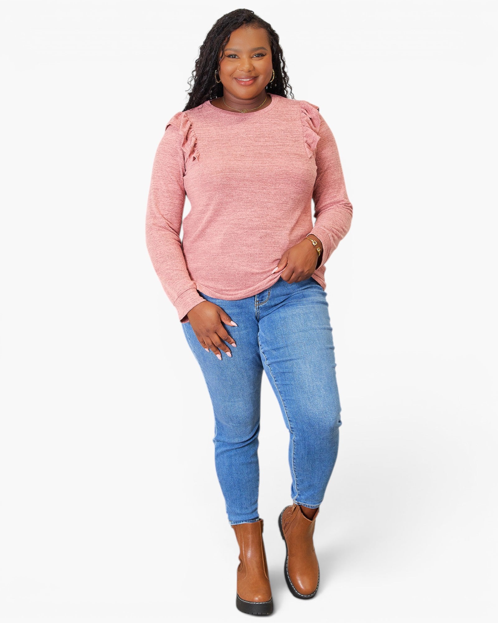 A woman modeling a pink long-sleeve knit top with ruffle details on the shoulders, paired with blue jeans and brown ankle boots, against a light gray studio background.