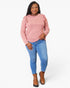 A woman modeling a pink long-sleeve knit top with ruffle details on the shoulders, paired with blue jeans and brown ankle boots, against a light gray studio background.
