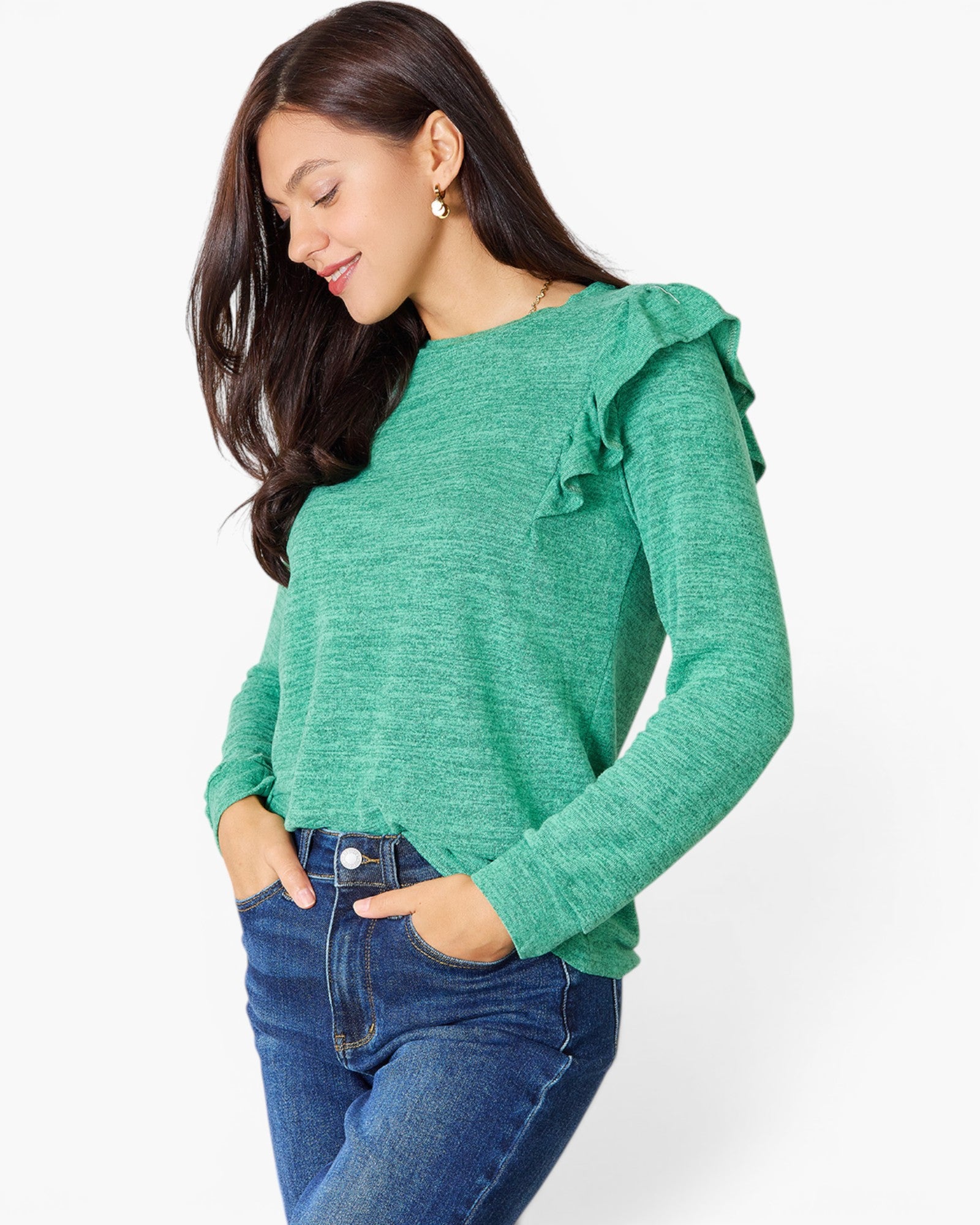 A woman wearing a green long-sleeve knit top with ruffle details on the shoulders, paired with blue jeans, photographed against a plain light gray background.