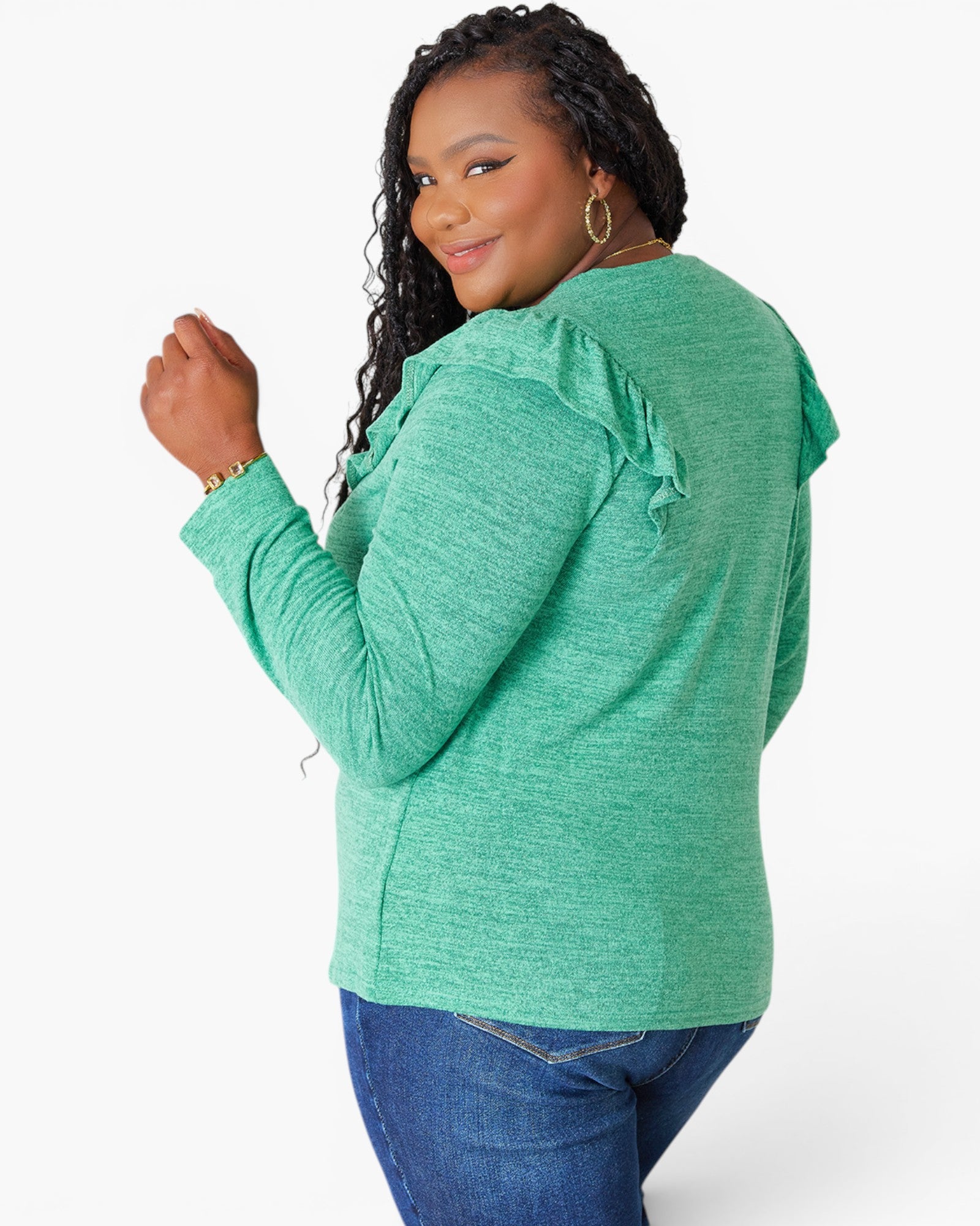 A woman wearing a heather green long-sleeve top with ruffle details on the shoulders, paired with blue jeans, photographed against a plain white studio background.