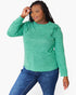 A woman modeling a green long-sleeve top with ruffle details on the shoulders, paired with blue jeans, against a plain white background.