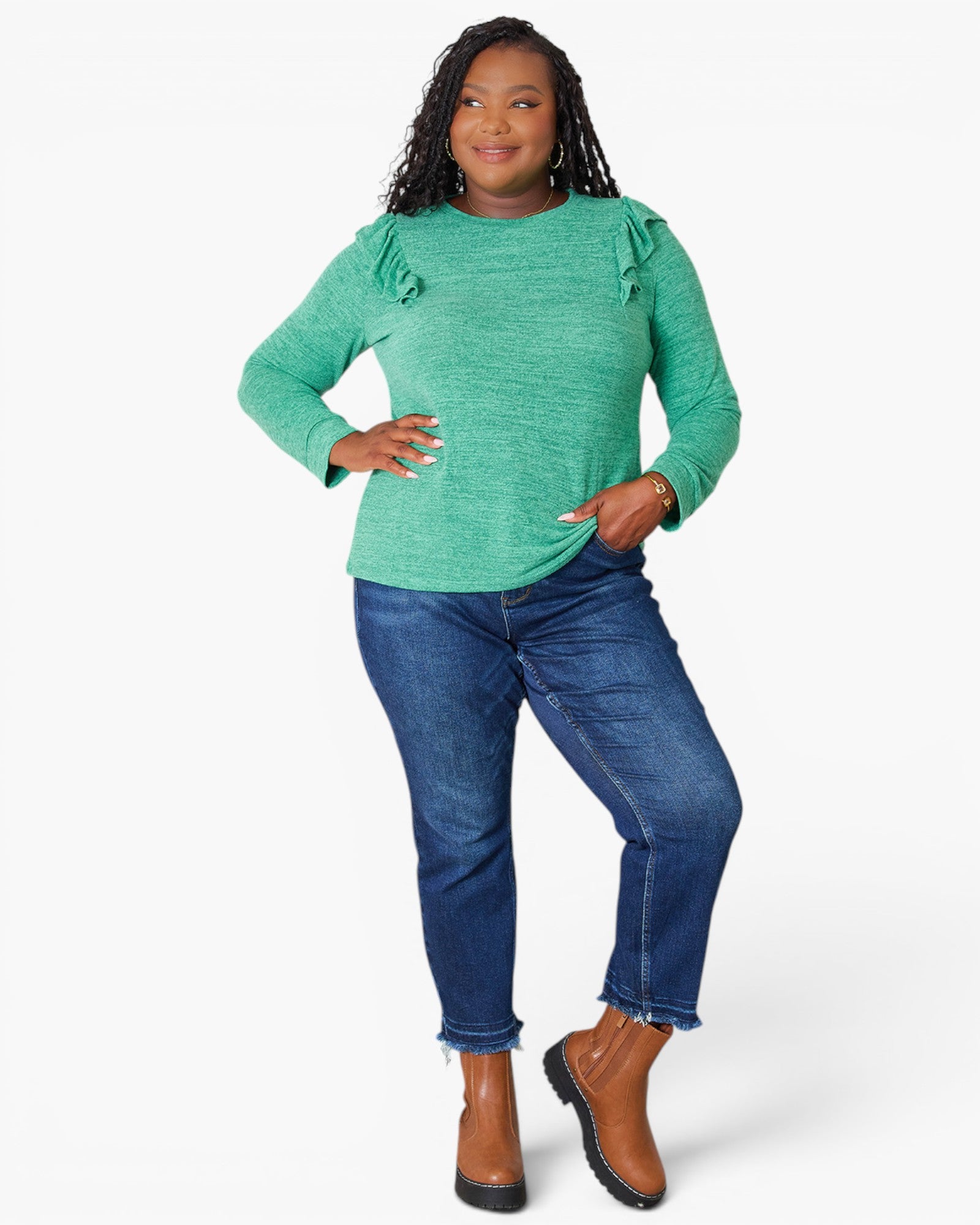 A woman modeling a green long-sleeve top with ruffle details on the shoulders, paired with blue jeans and brown ankle boots, against a plain white background.