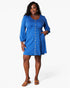 A woman modeling a blue long-sleeve mini dress with a button-down front, standing against a plain light gray studio background.