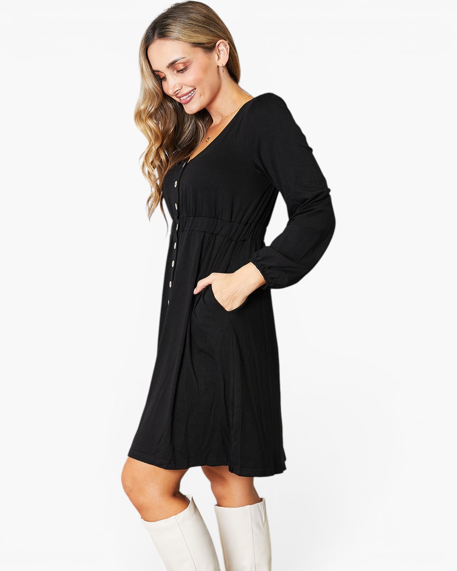A woman modeling a black long-sleeve V-neck dress with a button-front detail and side pockets, paired with white knee-high boots, against a plain white studio background.