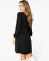 A woman wearing a black long-sleeve dress with a gathered waist, shown from a three-quarter back angle against a plain white studio background.