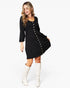 A woman modeling a black knee-length dress with three-quarter sleeves, a round neckline, and decorative gold-tone buttons down the front, paired with white knee-high boots against a light gray studio background.