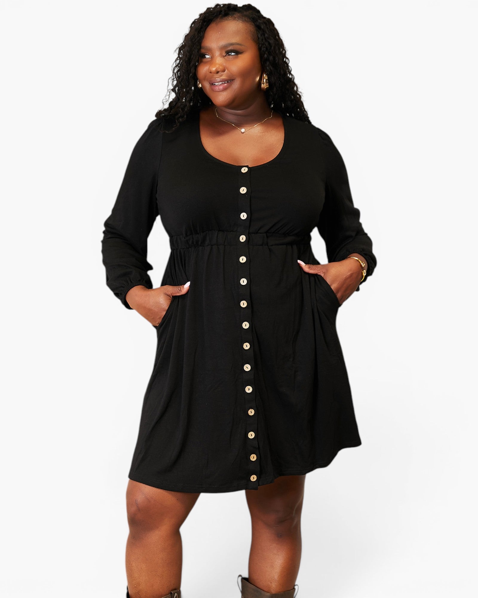 A plus-size woman modeling a black long-sleeve mini dress with a button-down front and side pockets, set against a plain light gray studio background.