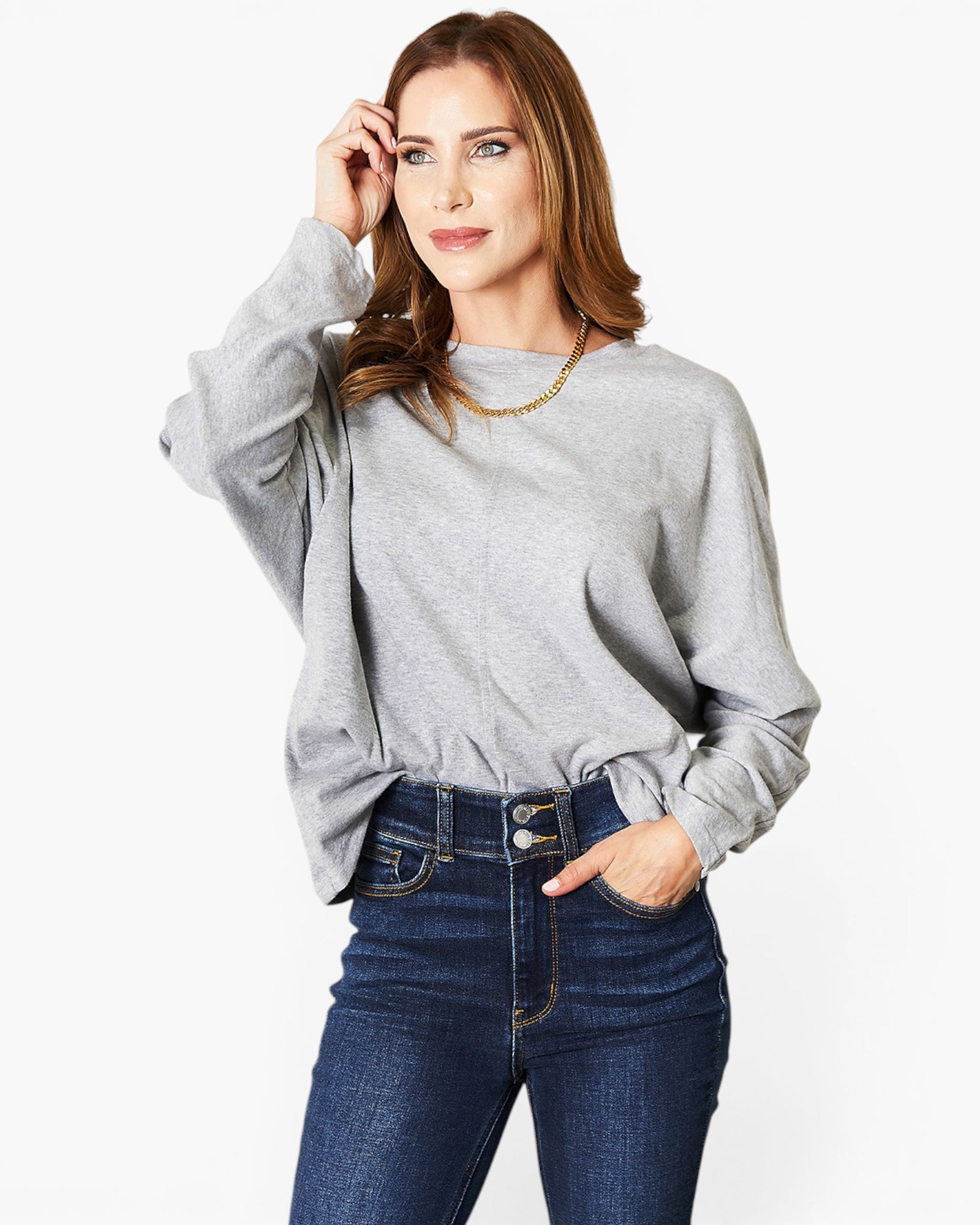 A woman wearing a light gray oversized long-sleeve sweatshirt and dark blue jeans, standing against a plain white studio background.