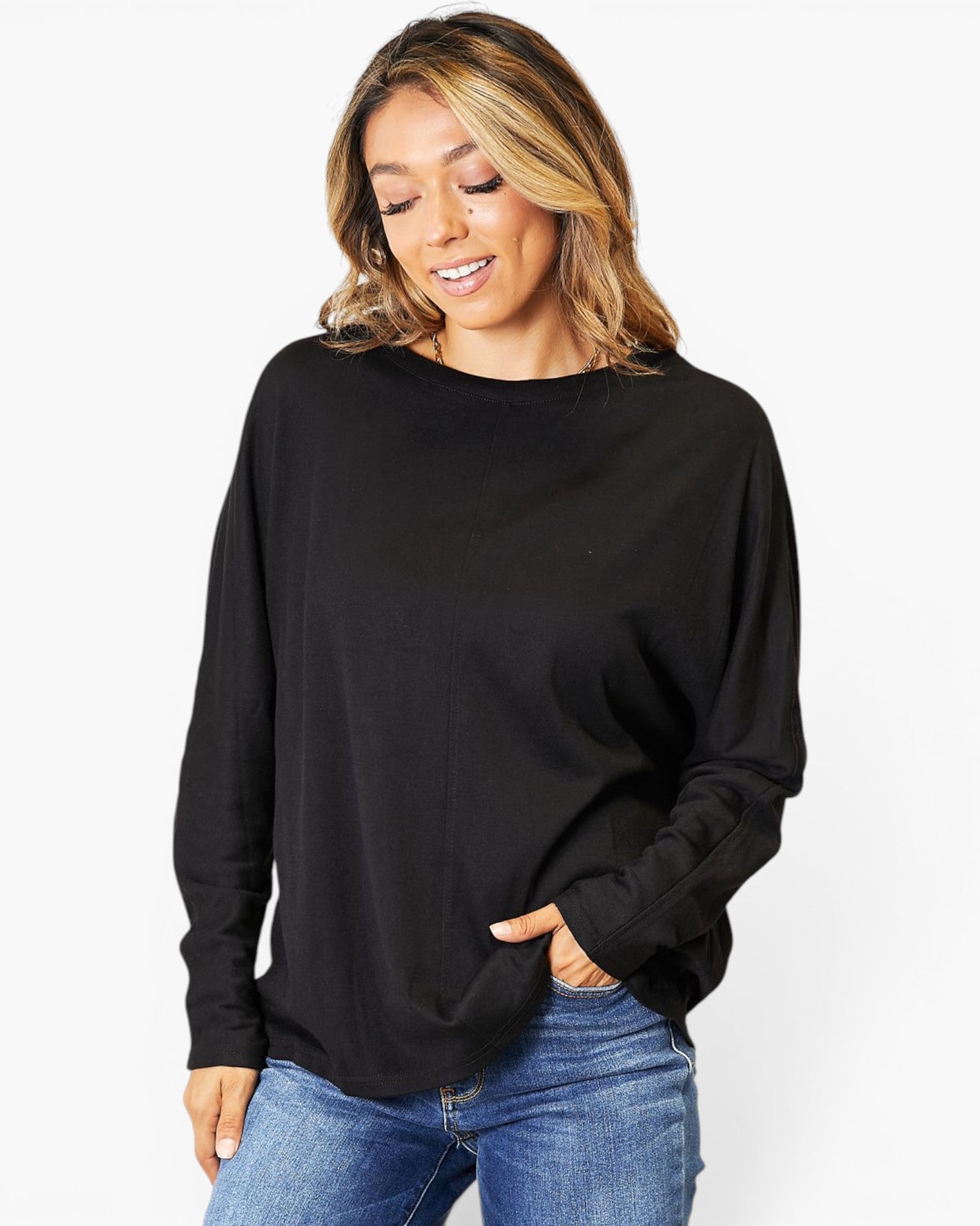 A woman wearing a black long-sleeve knit top with a wide boat neckline and blue jeans, standing against a plain white studio background.