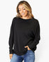 A woman wearing a black long-sleeve knit top with a wide boat neckline and blue jeans, standing against a plain white studio background.