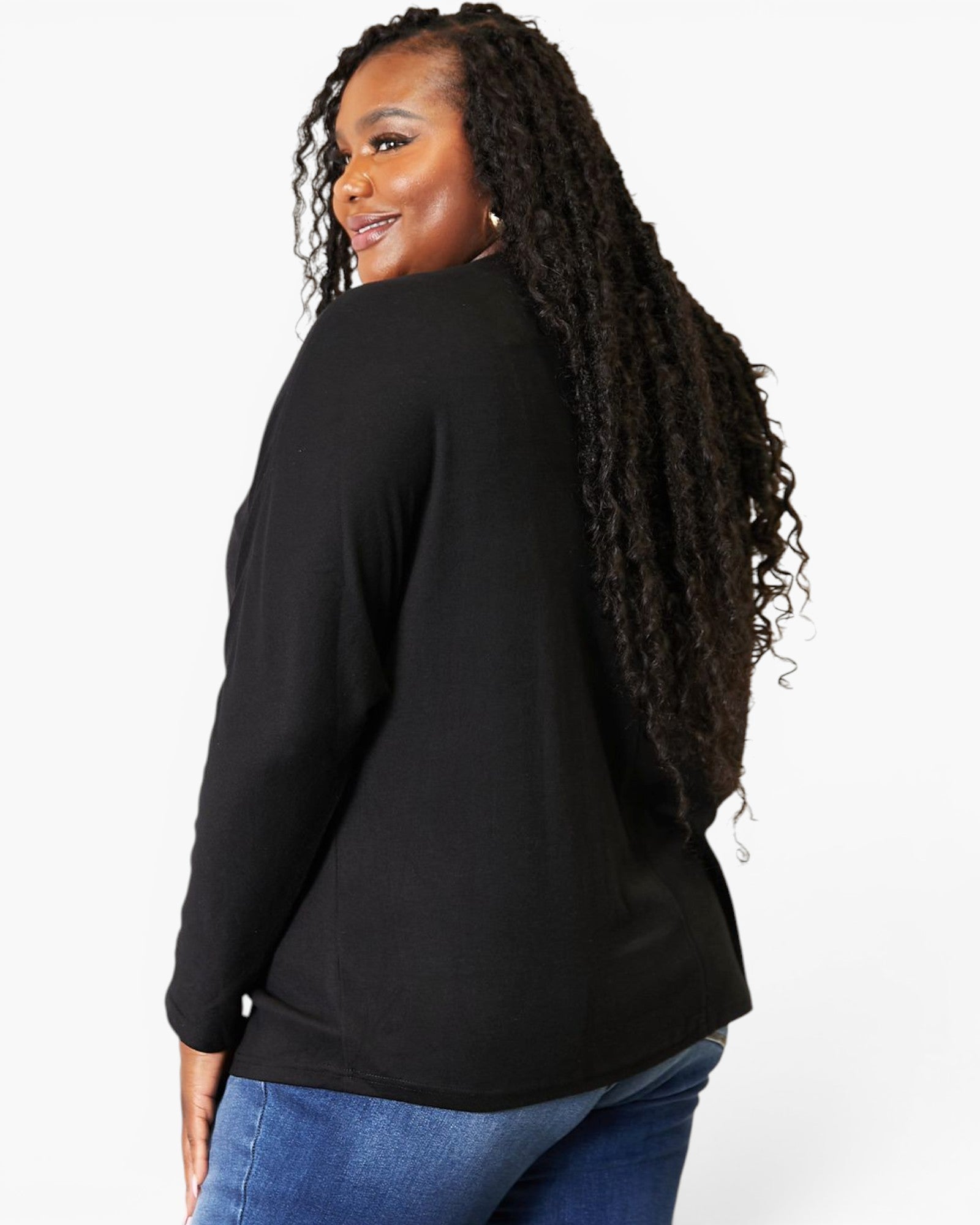 A woman with long, curly black hair is shown from a side-back angle wearing a black long-sleeve top and blue jeans against a plain white studio background.