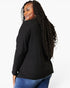 A woman with long, curly black hair is shown from a side-back angle wearing a black long-sleeve top and blue jeans against a plain white studio background.