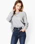 A woman wearing a light gray long-sleeve crewneck sweatshirt and dark blue jeans, posing against a plain white studio background.