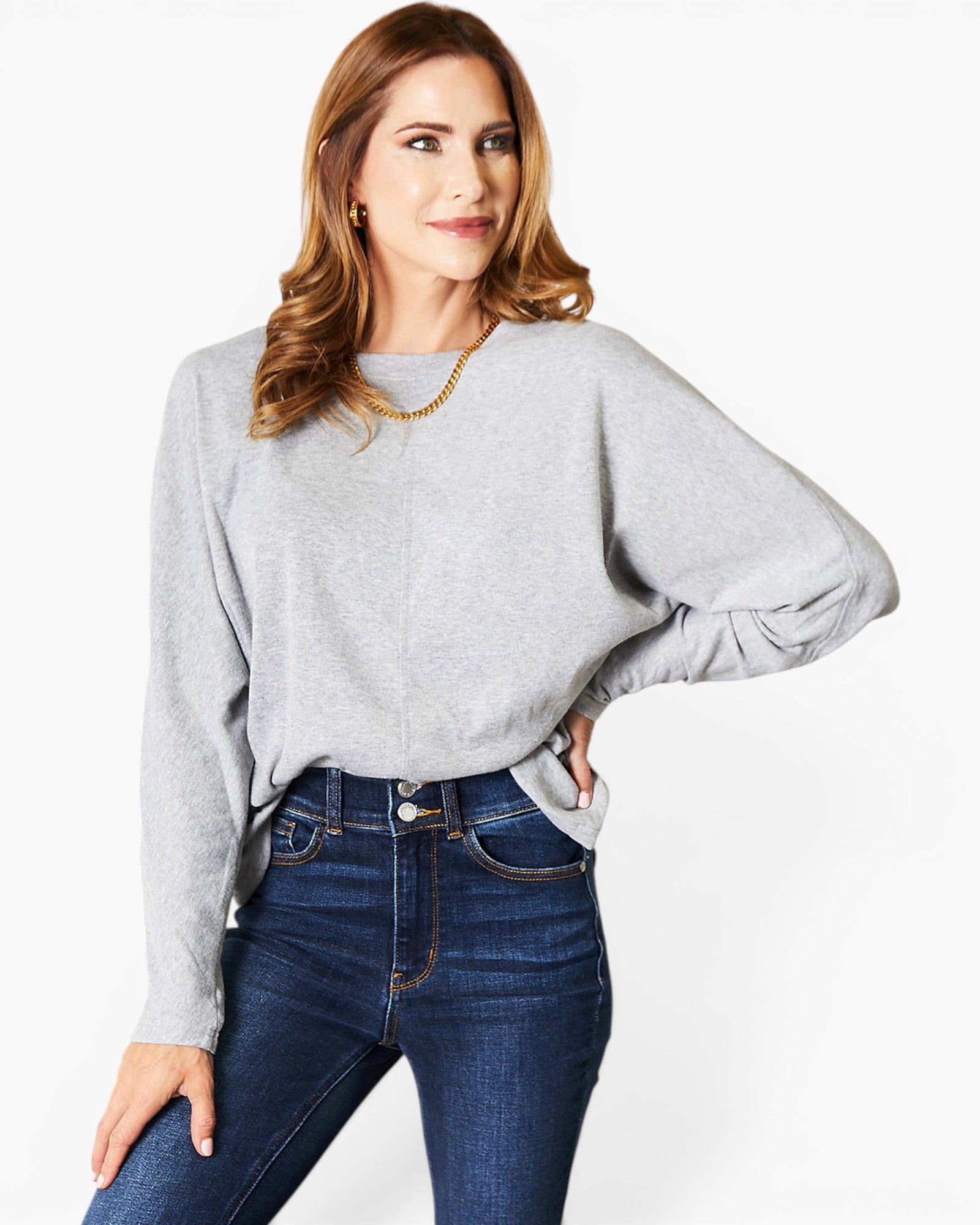 A woman wearing a light gray long-sleeve knit top with balloon sleeves and dark blue jeans, standing against a plain white studio background.