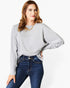A woman wearing a light gray long-sleeve knit top with balloon sleeves and dark blue jeans, standing against a plain white studio background.
