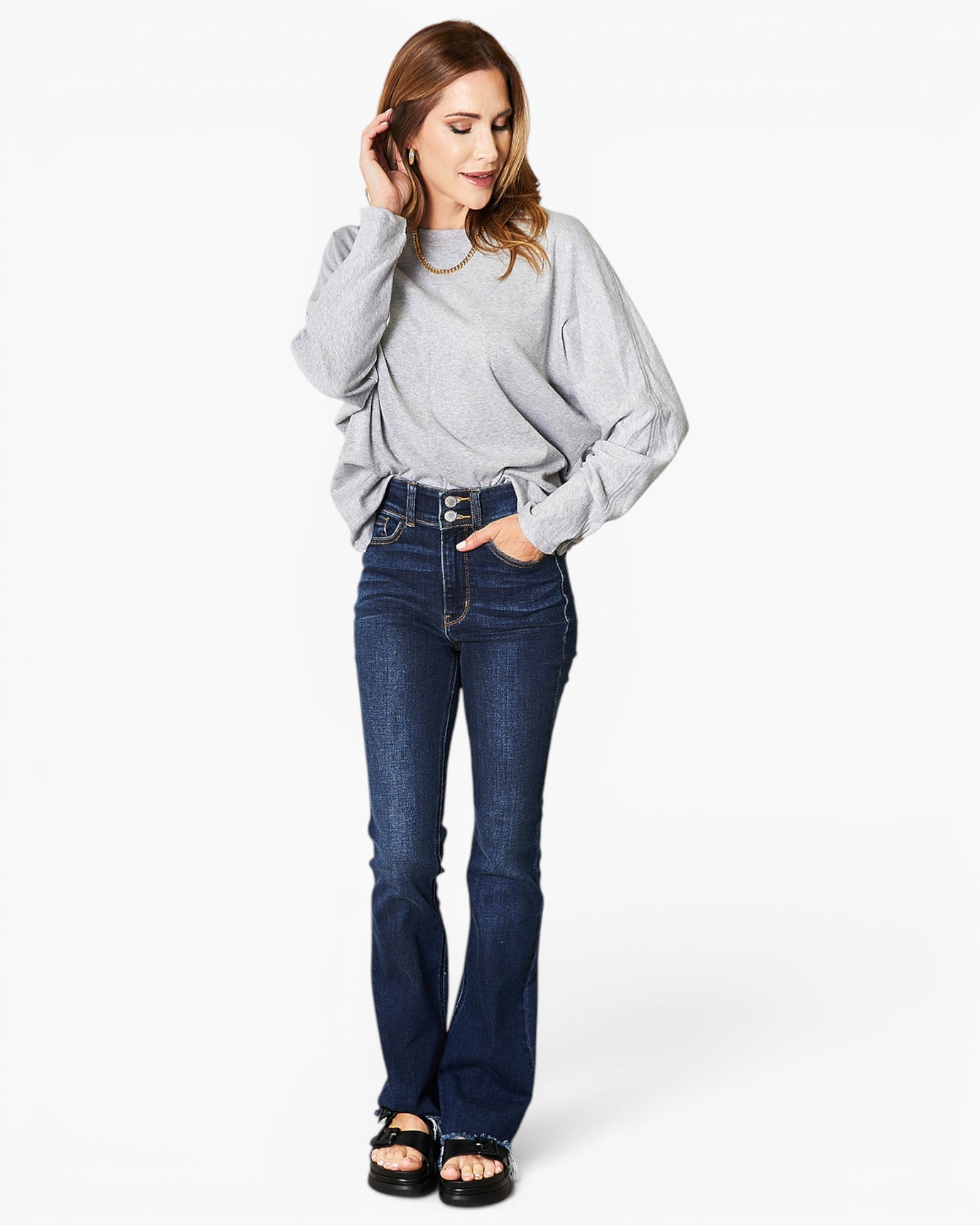 A woman modeling a light gray long-sleeve top and dark blue flared jeans with black sandals, standing against a plain white studio background.