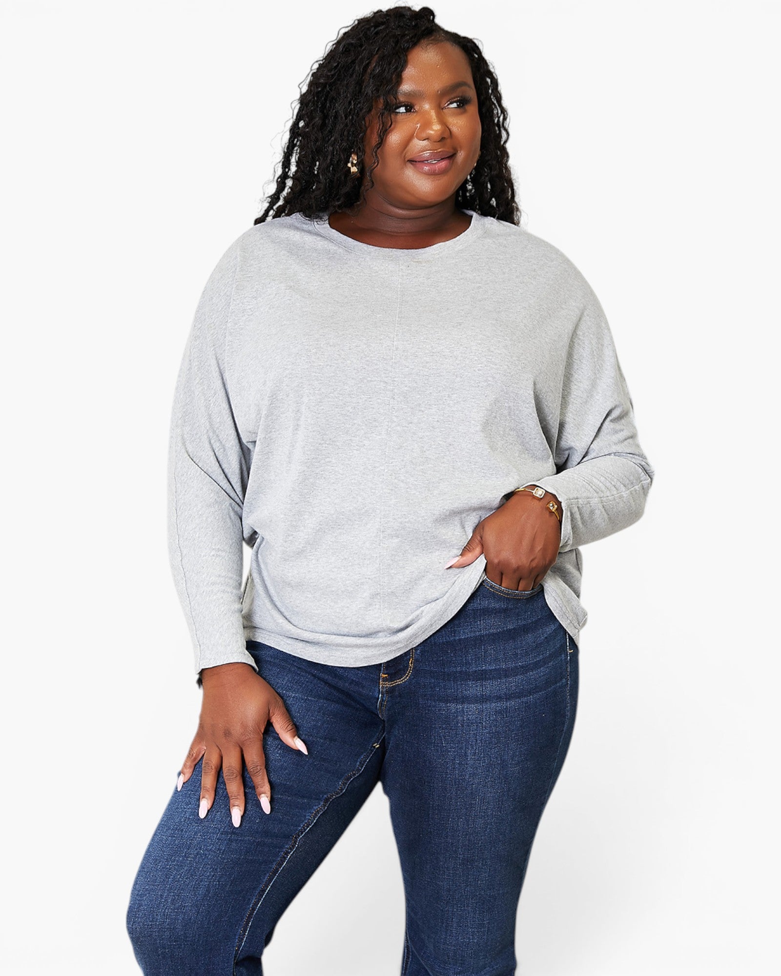 A woman modeling a light gray long-sleeve knit top with a relaxed fit, paired with dark blue jeans, against a plain white studio background.
