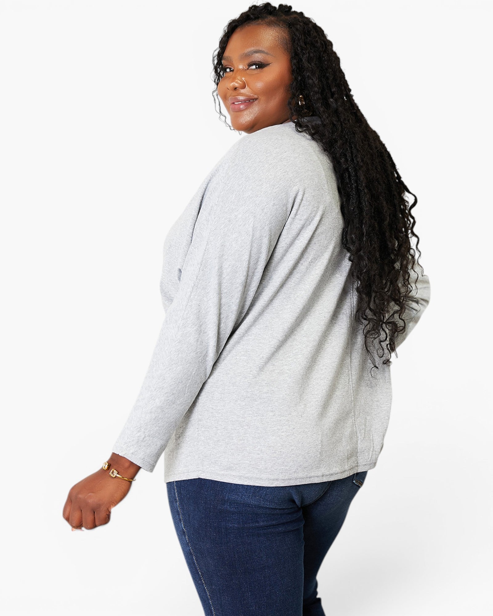 A woman with long, curly black hair is shown from a side-back angle wearing a light gray heathered long-sleeve top and dark blue jeans against a plain white studio background.
