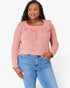 A woman modeling a dusty pink long-sleeve top with ruffle details on the shoulders and a square neckline, paired with blue jeans, against a plain white background.