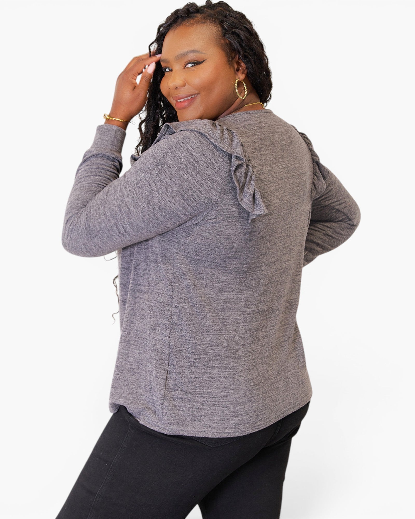 A woman modeling a heather gray long-sleeve top from the back, featuring ruffled shoulder details and a relaxed fit, paired with black pants against a plain white background.