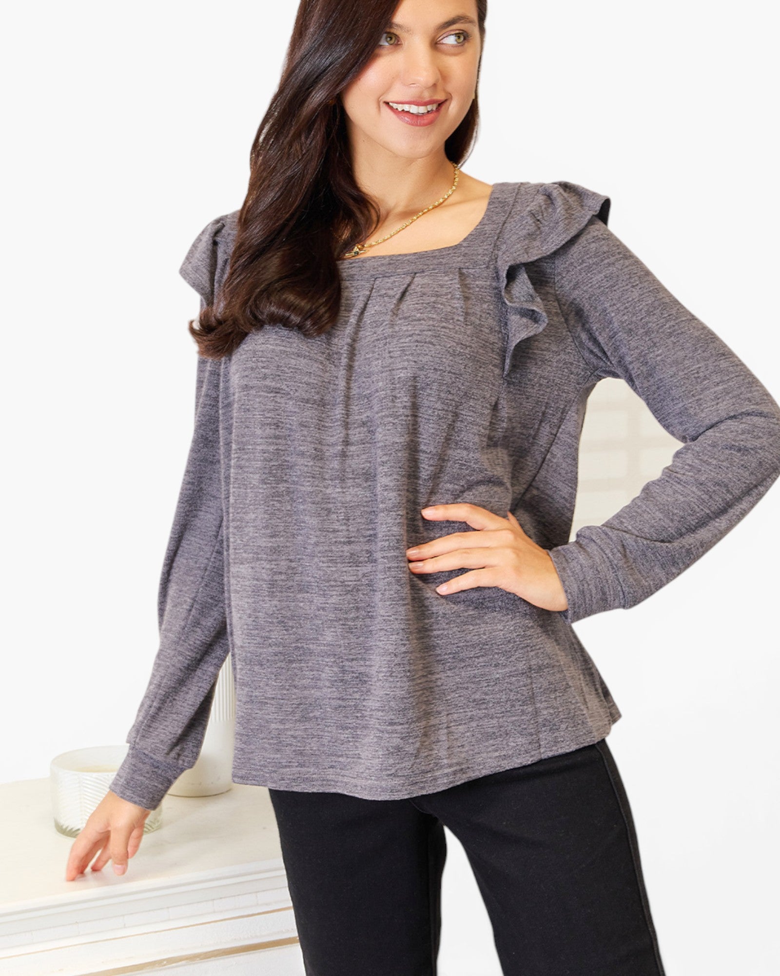 A woman wearing a heather gray long-sleeve blouse with ruffled shoulder details and a square neckline, paired with black pants, standing against a white background.