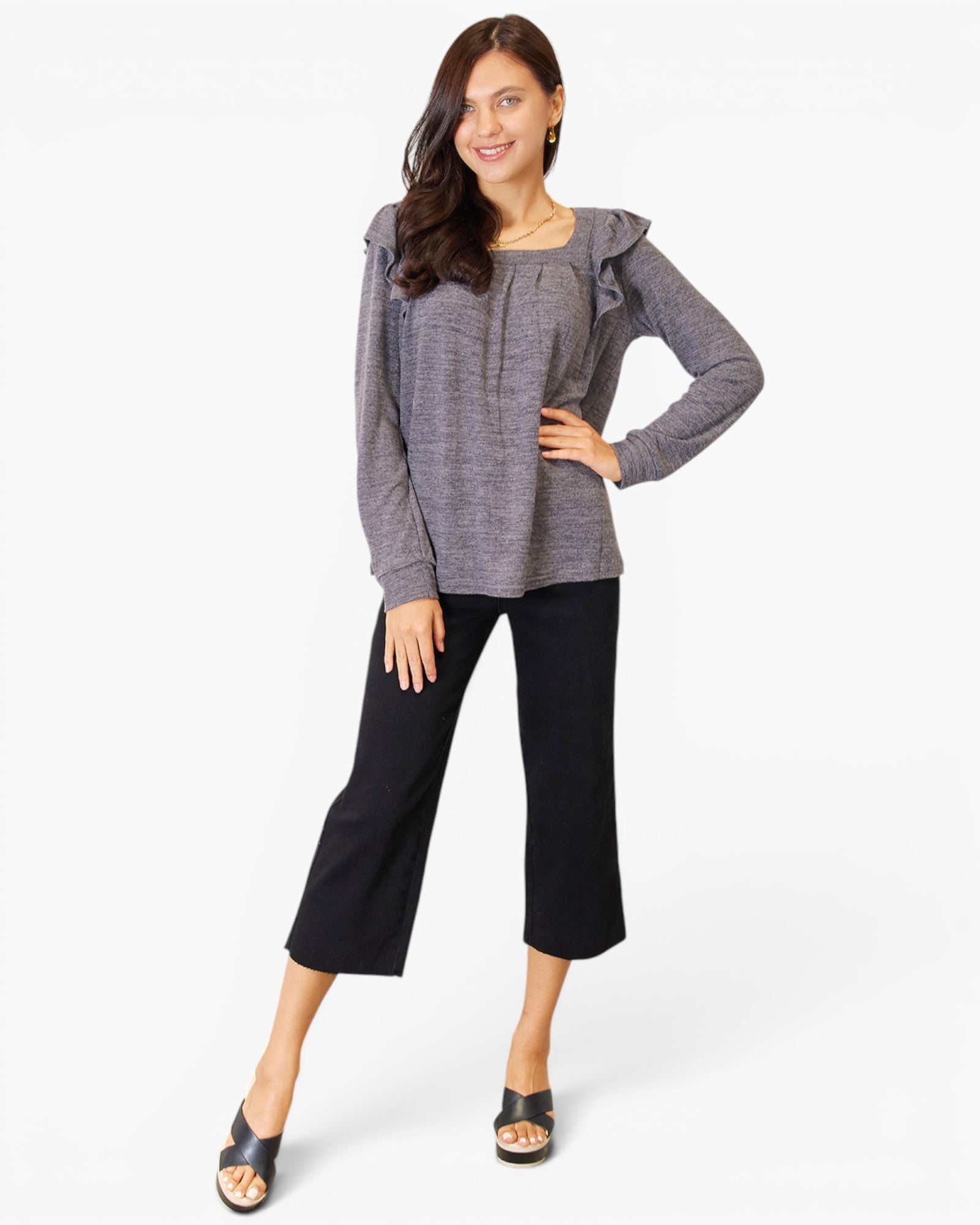 A woman modeling a gray long-sleeve top with ruffled shoulder details and black cropped pants, standing against a plain white studio background.