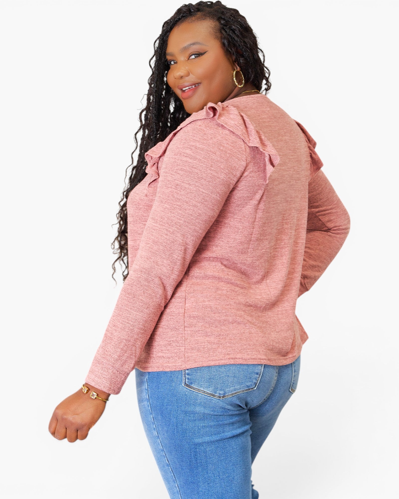 A woman wearing a heathered pink long-sleeve top with ruffled shoulder details and blue jeans, photographed against a plain light gray studio background.