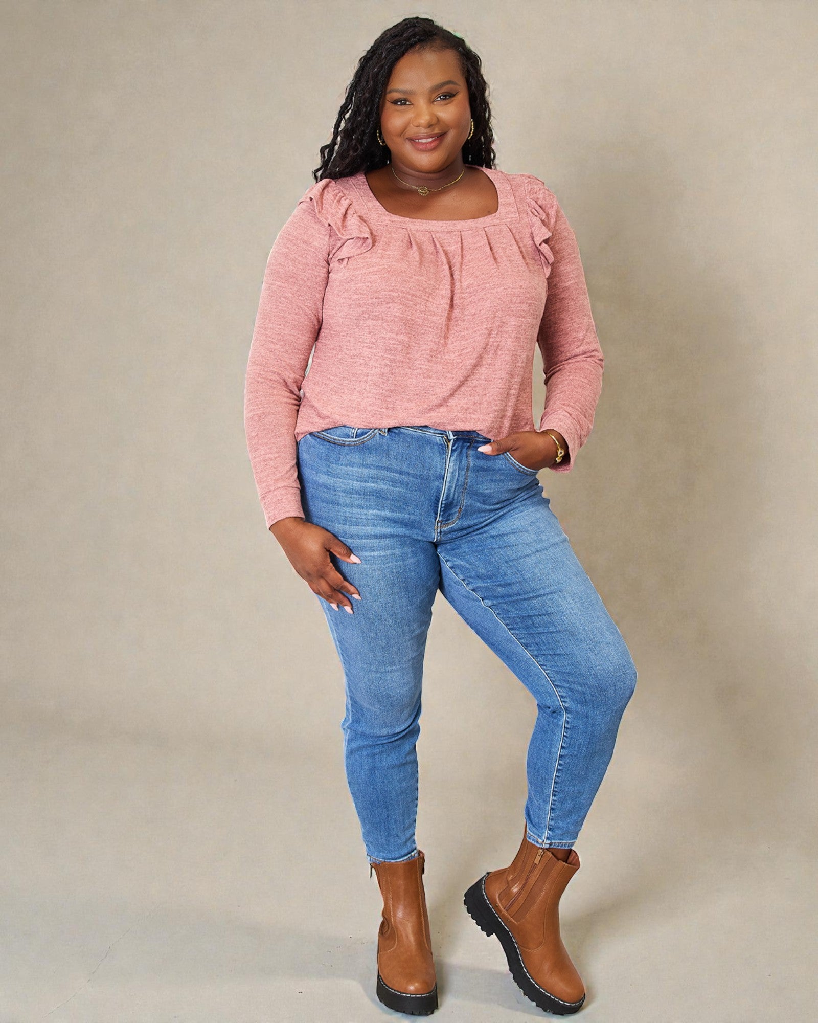 Woman wearing a pink top and blue jeans with brown boots against a beige background