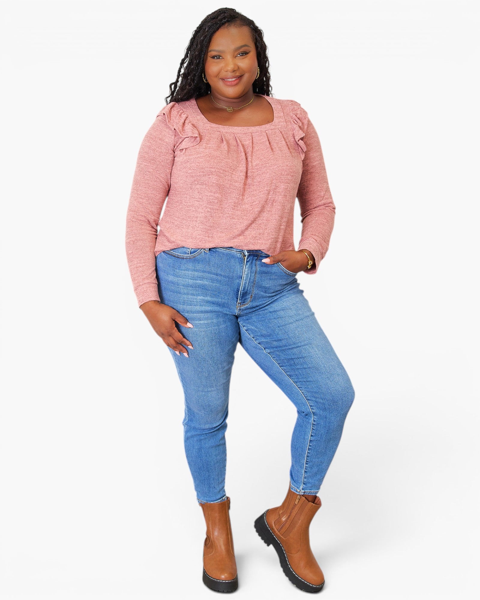 A woman modeling a pink long-sleeve top with ruffle details on the shoulders and a square neckline, paired with blue skinny jeans and brown ankle boots, against a light gray studio background.