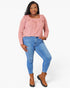 A woman modeling a pink long-sleeve top with ruffle details on the shoulders and a square neckline, paired with blue skinny jeans and brown ankle boots, against a light gray studio background.