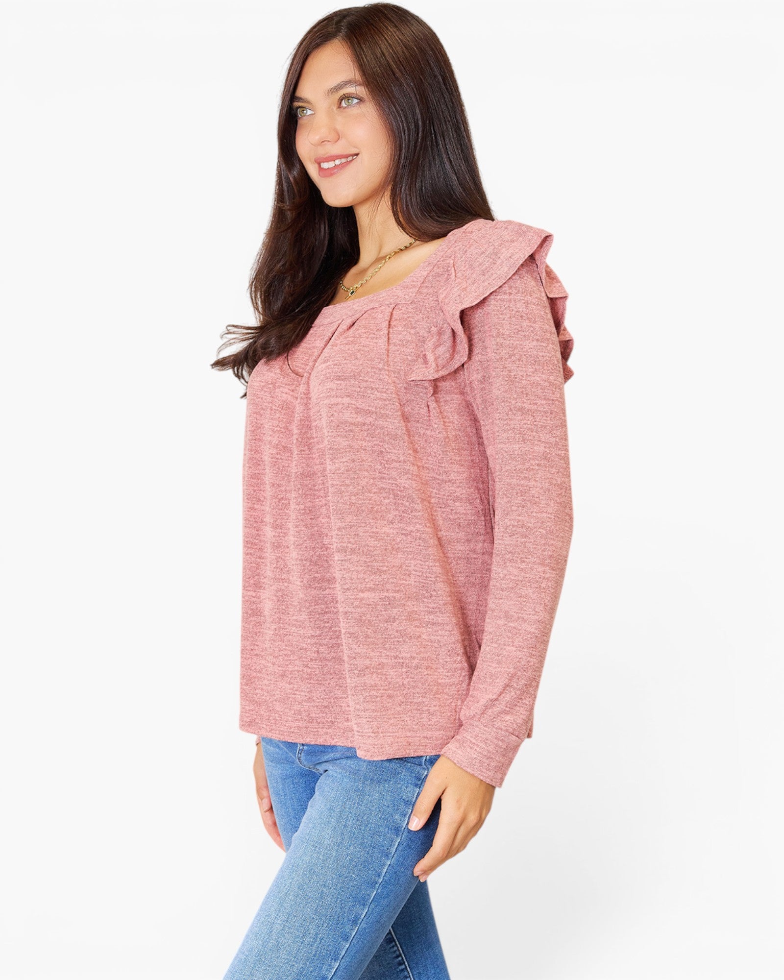 A woman wearing a heathered pink long-sleeve top with ruffled shoulder details and blue jeans, photographed against a plain white studio background.