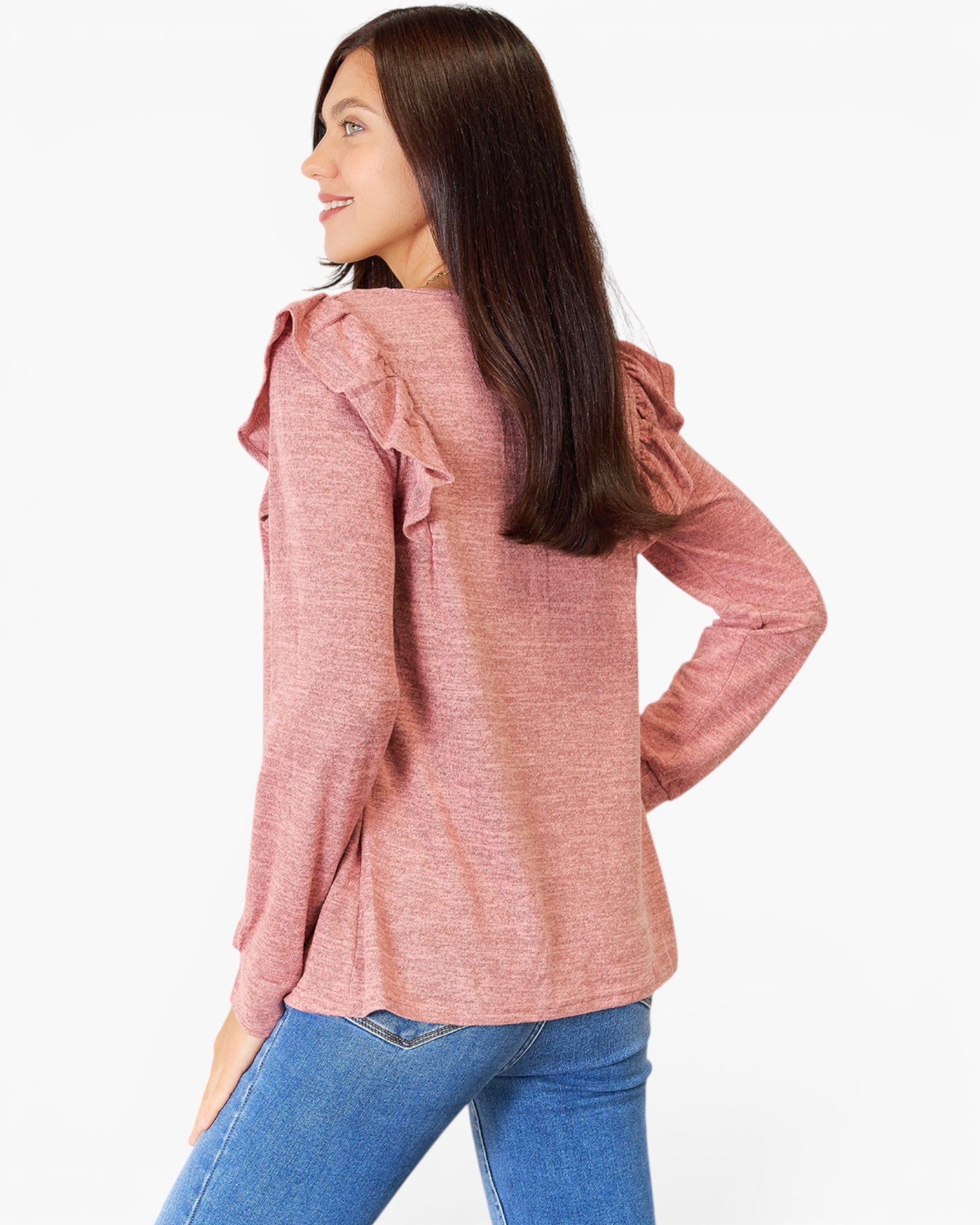 A woman wearing a dusty rose pink long-sleeve top with ruffle details on the shoulders, paired with blue jeans, shown from a side-back angle against a plain white background.
