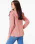 A woman wearing a dusty rose pink long-sleeve top with ruffle details on the shoulders, paired with blue jeans, shown from a side-back angle against a plain white background.