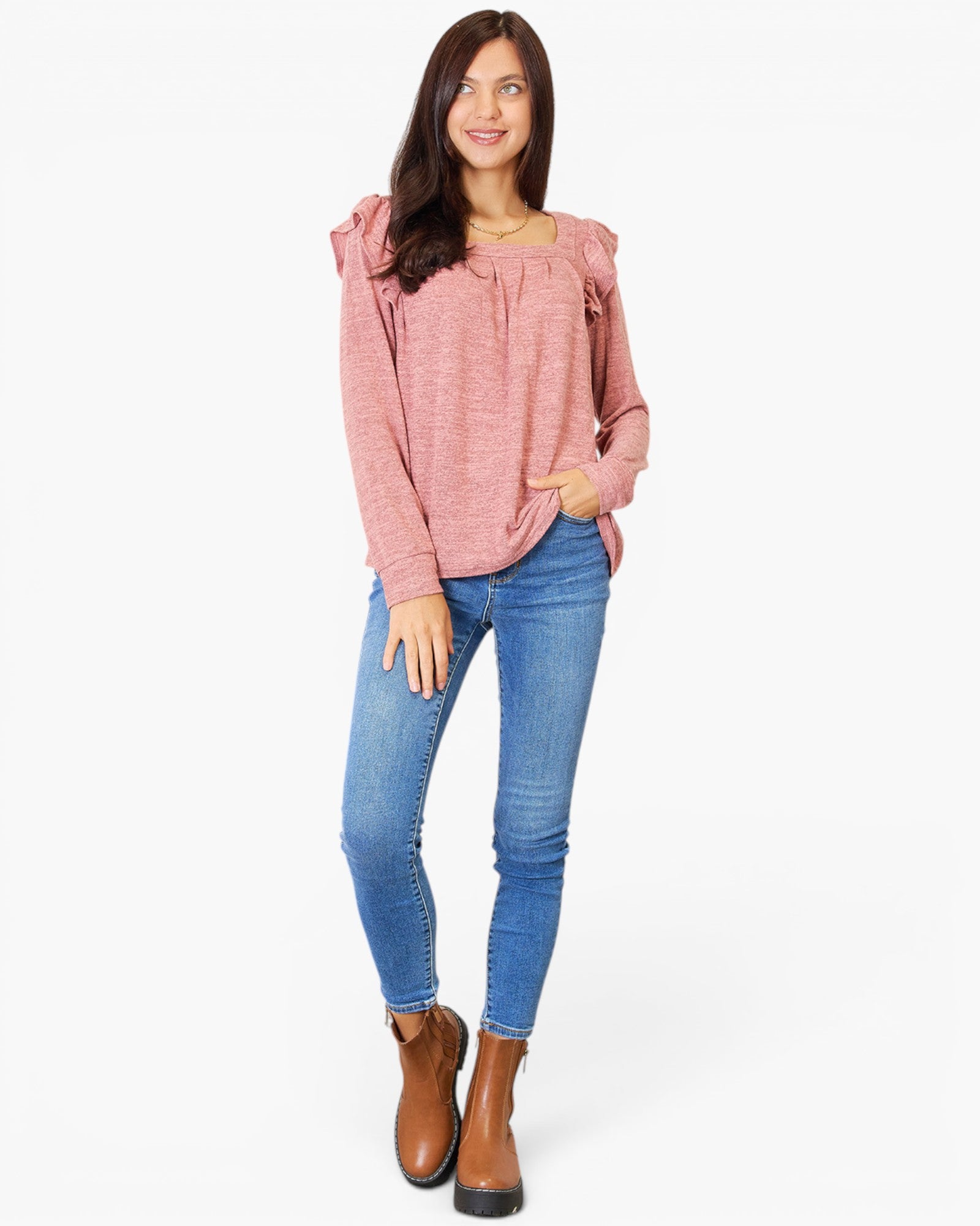 A woman modeling a dusty pink long-sleeve top with ruffled shoulder details, paired with blue skinny jeans and brown ankle boots, against a plain white background.