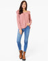 A woman modeling a dusty pink long-sleeve top with ruffled shoulder details, paired with blue skinny jeans and brown ankle boots, against a plain white background.