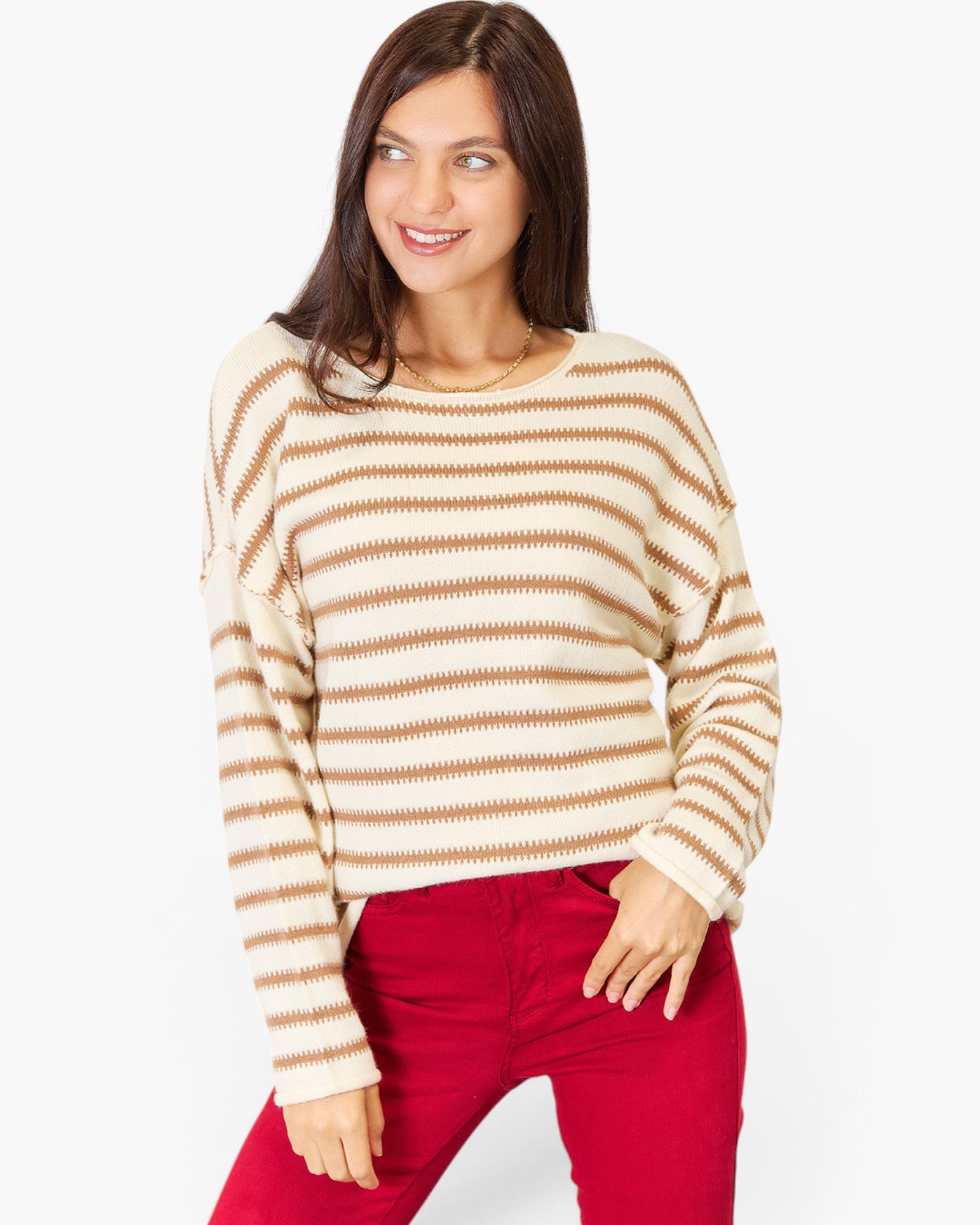 A woman wearing a cream and tan horizontally striped knit sweater with red pants, standing against a plain light gray studio background.