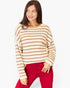 A woman wearing a cream and tan horizontally striped knit sweater with red pants, standing against a plain light gray studio background.