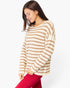 A woman wearing a cream and tan striped knit sweater and bright pink pants, shown from the waist up against a plain light gray studio background.