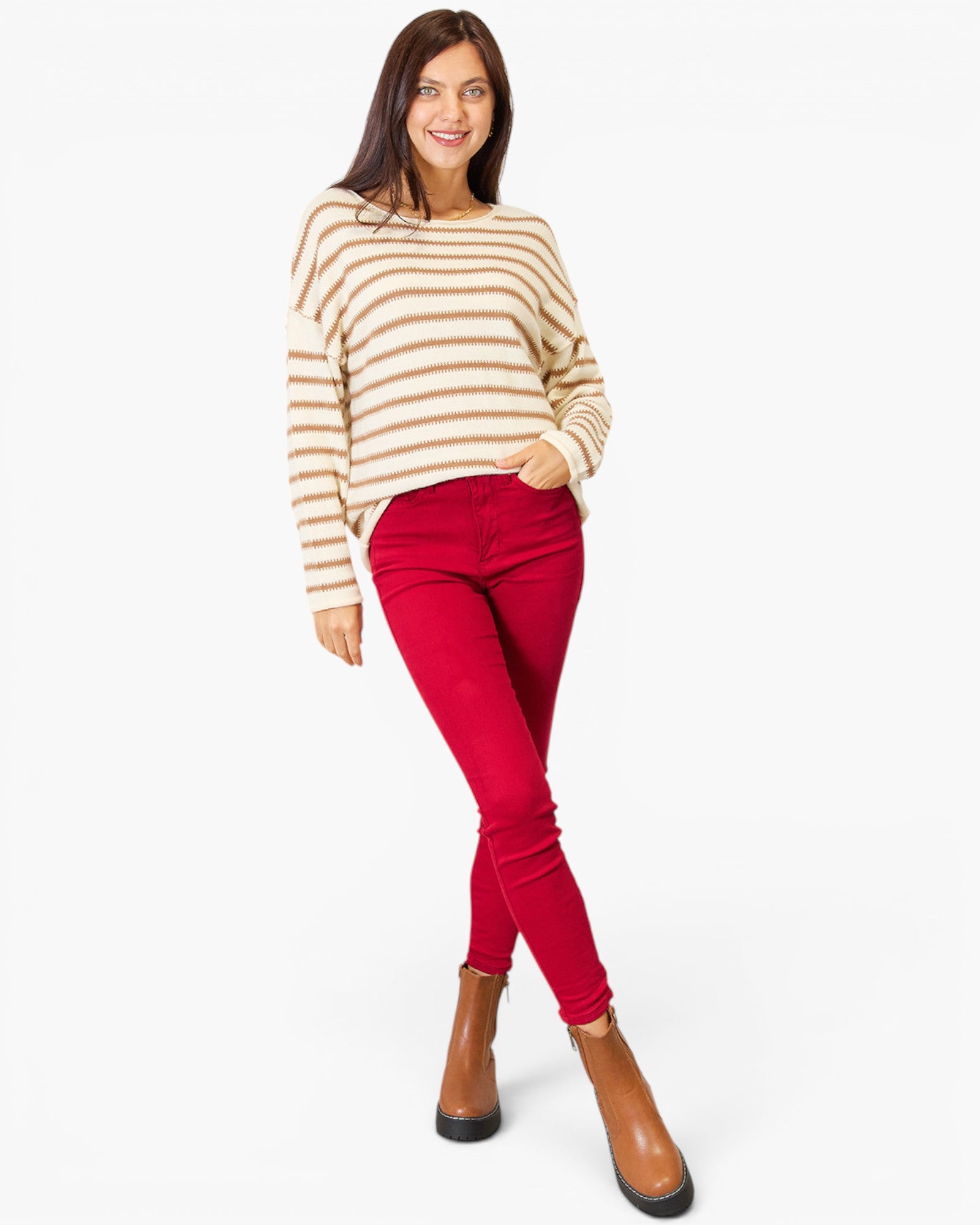 A woman modeling a cream and tan striped knit sweater paired with bright red skinny jeans and brown ankle boots, photographed against a plain white studio background.