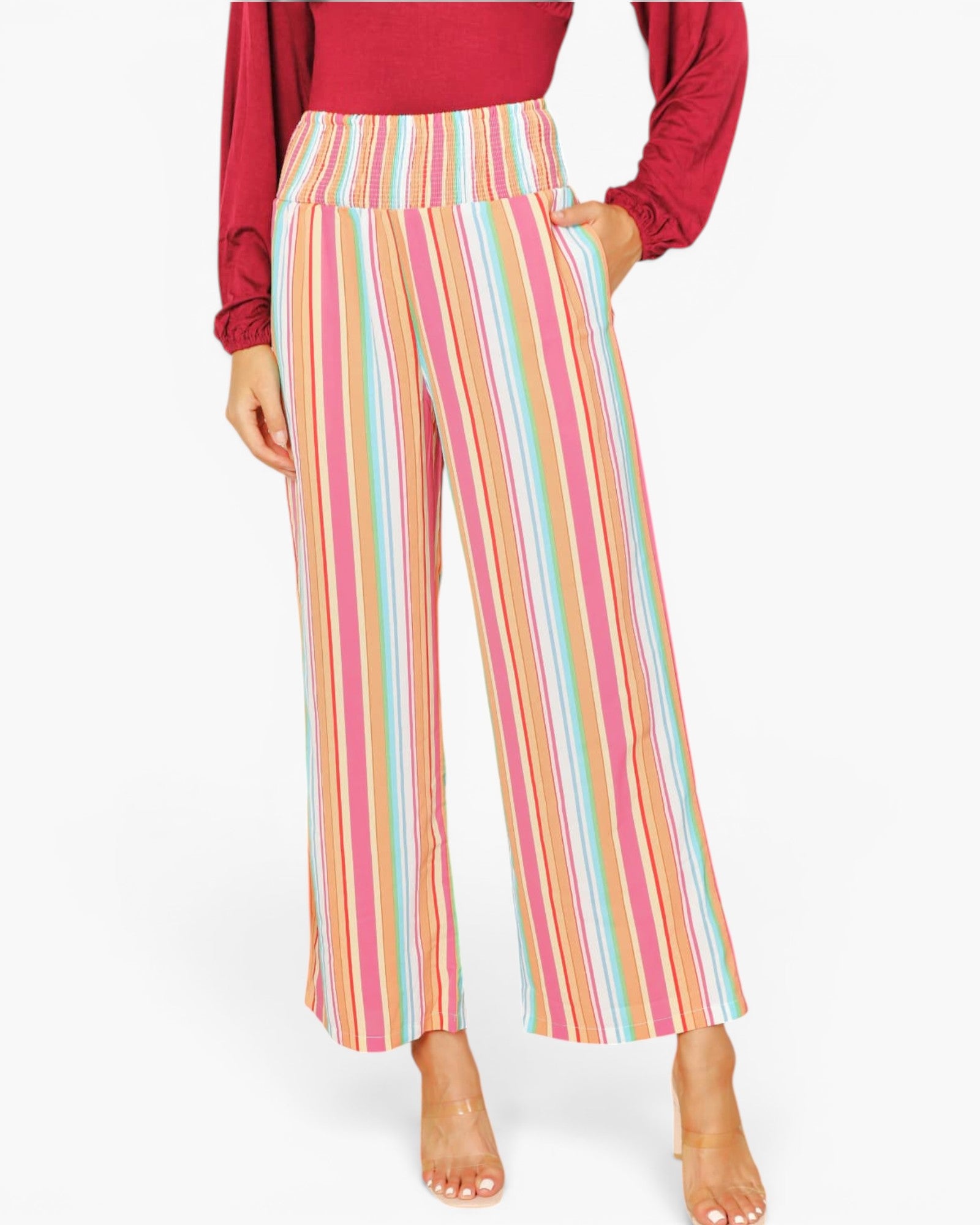 A model wearing high-waisted, wide-leg cropped pants with vertical pastel stripes in pink, orange, yellow, green, and blue, paired with a red long-sleeve top and clear high-heeled sandals.
