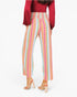 A model from the back wearing high-waisted, wide-leg cropped pants with vertical pastel stripes in pink, orange, yellow, green, and blue, paired with a red long-sleeve top and beige block-heel sandals.